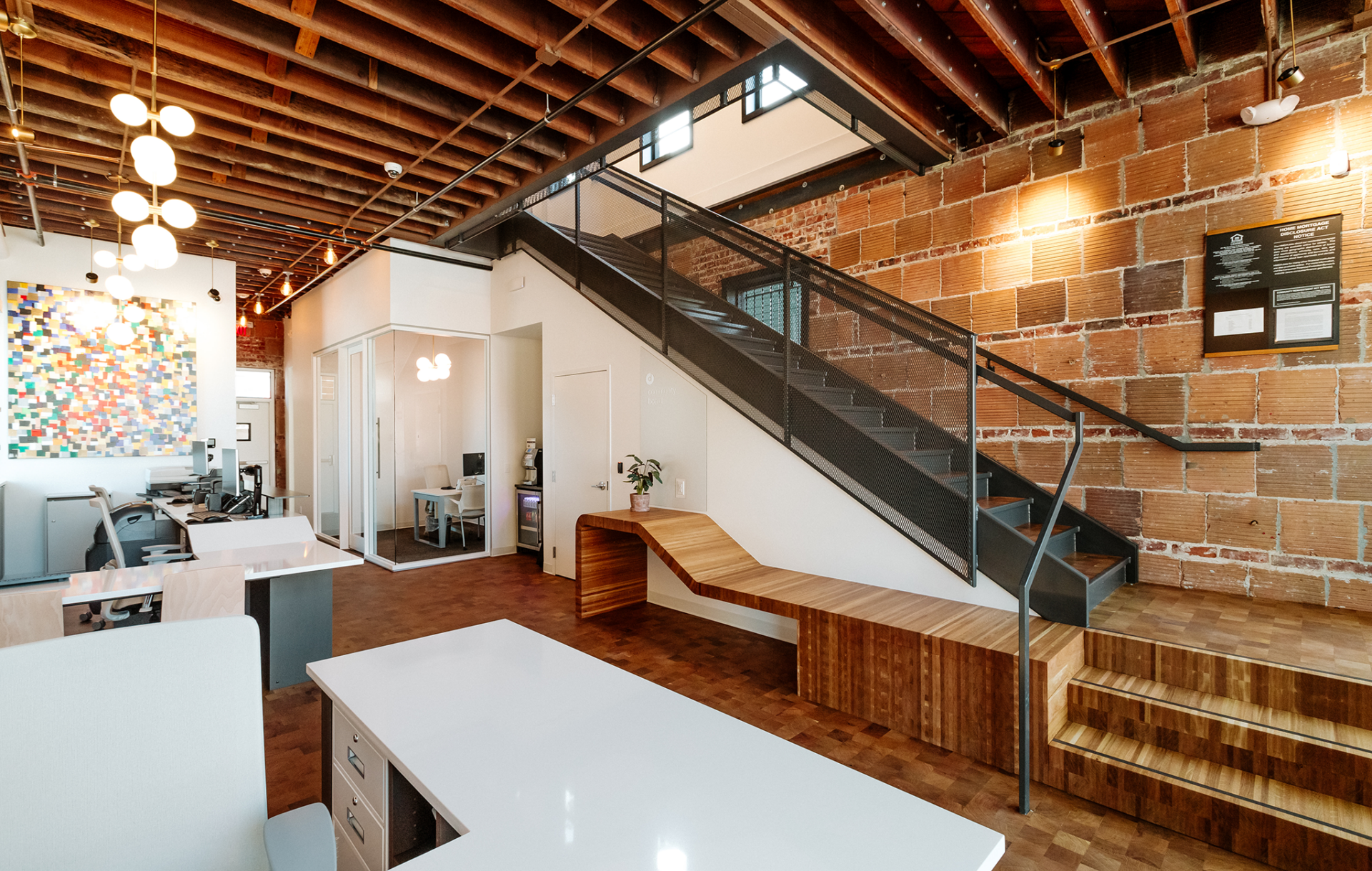 A bank lobby. Teller stations to the left and a staircase to the right. Brick walls, wood floors, and modern furniture.