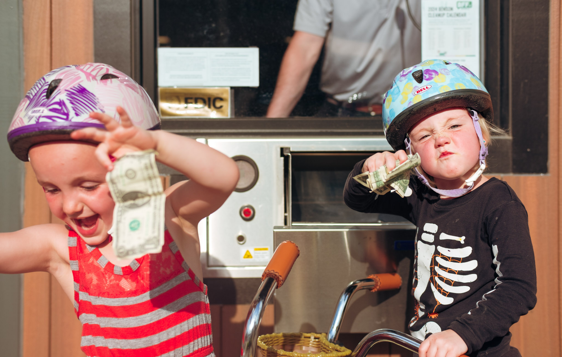 Two kids celebrate with dollar bills in a bank drive-thru.